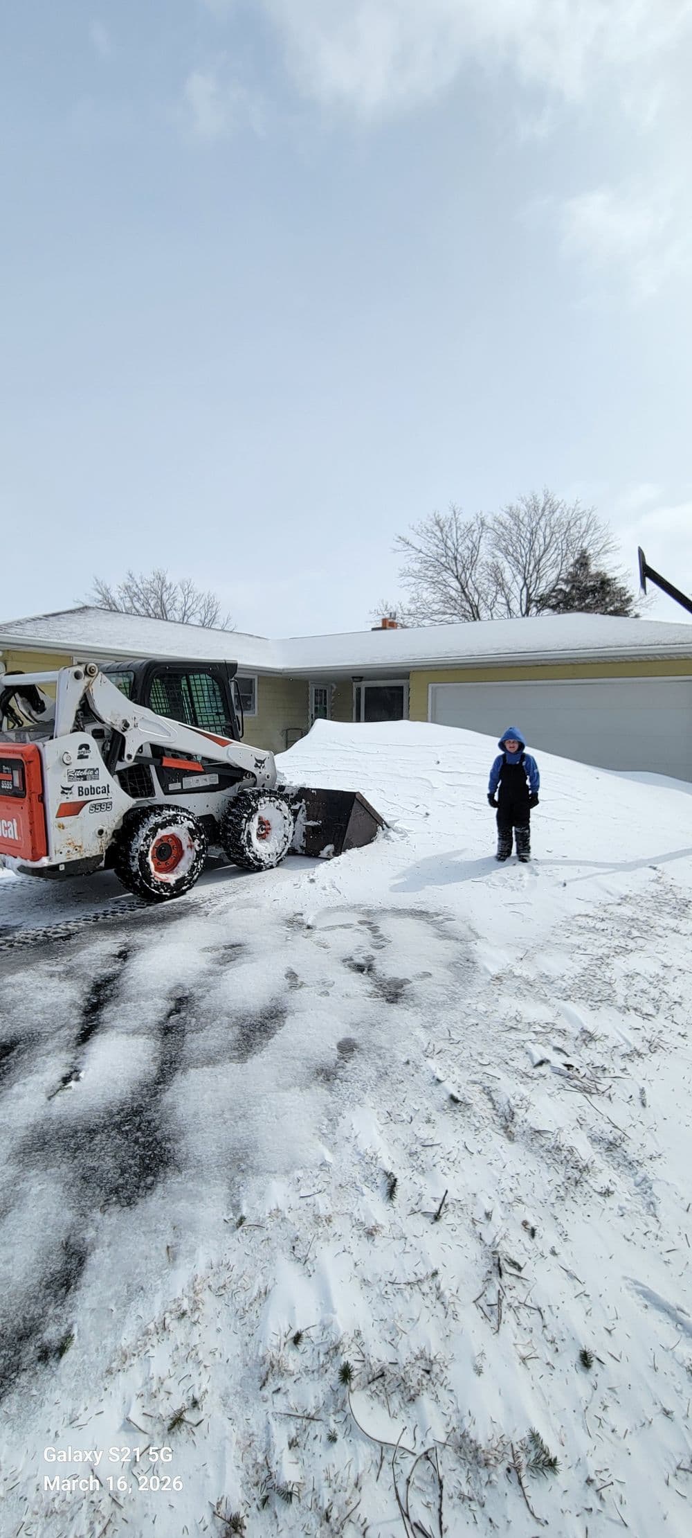 Child standing beside a snow plow clearing snow from a residential driveway.