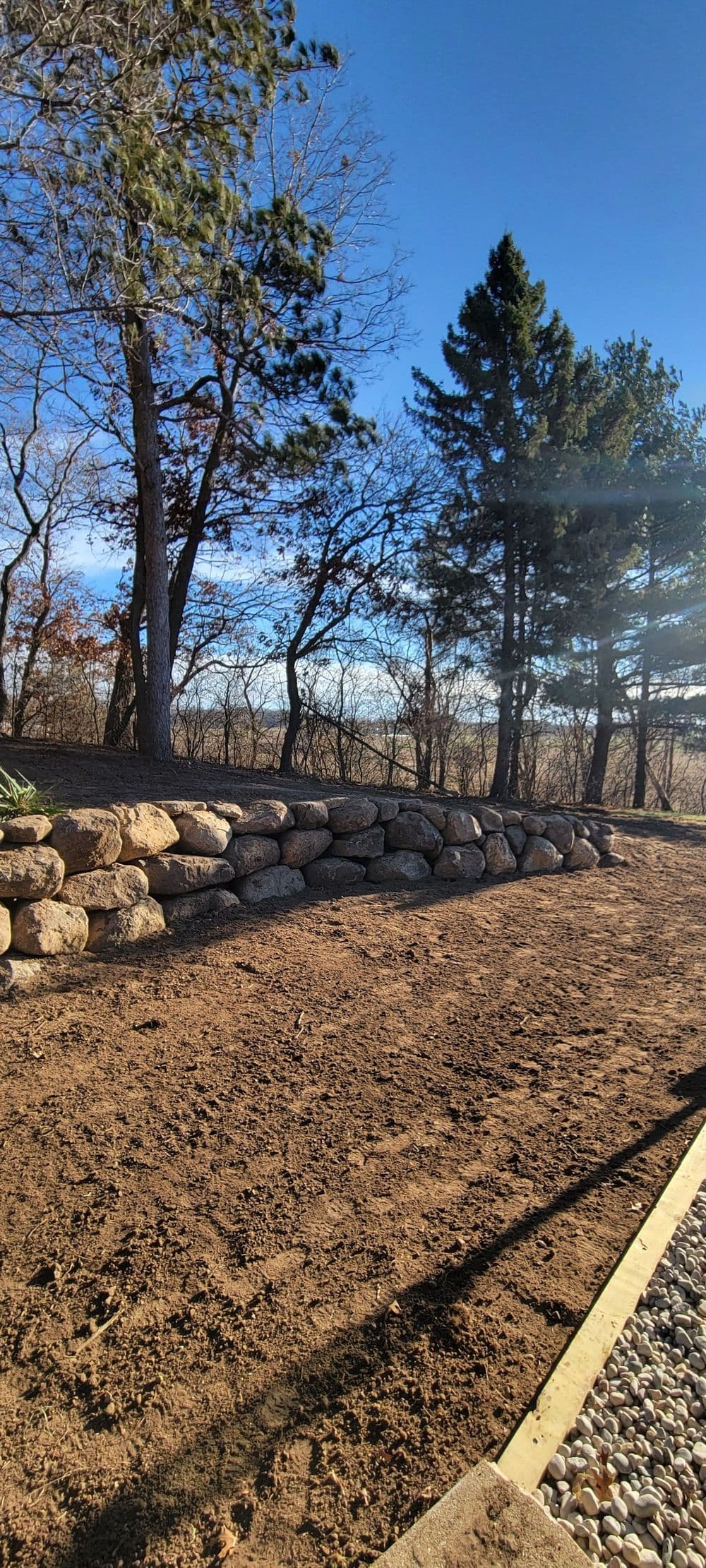 Stone retaining wall in a sunlit outdoor landscape with trees and clear blue sky.